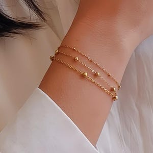 Könnte beinhalten: Drei goldene Kettenarmbänder mit kleinen runden Perlen. Die Armbänder sind übereinander am Handgelenk einer Person gelegt.