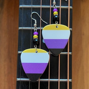 May include: Non-binary pride flag earrings. Each earring features a guitar pick shape with the non-binary flag colors: yellow, white, purple, and black. The earrings have silver hooks and are adorned with purple, yellow, and black beads.