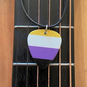 May include: A guitar pick pendant with the non-binary pride flag design. The pick features horizontal stripes in gold, white, purple, and black. It hangs from a black cord necklace, resting on the fretboard of a guitar.