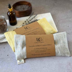 Può includere: Due cuscini comfort profumati alla lavanda e al basilico con un'etichetta marrone che recita "Lavender & Basil Comfort Pillow Soothing & Relaxing Use as an eye pillow or as a compress for neck & other body aches and pains Weighted with organic flax seeds". I cuscini sono realizzati in lino grigio chiaro e giallo.