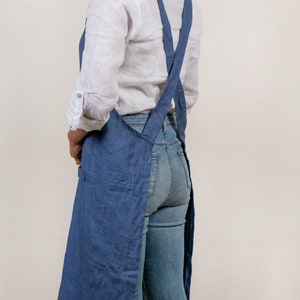 May include: A blue linen apron with adjustable criss-cross straps. The apron is long and has a pocket on the front.