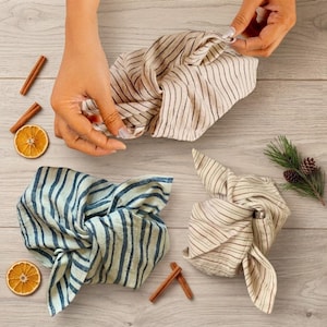 Peut inclure: Trois emballages en tissu noués, deux avec des rayures verticales beige et marron et un avec des rayures verticales bleu clair et bleu foncé. Les emballages sont disposés avec des tranches d'orange séchées, des bâtons de cannelle et un brin de pin.