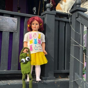 May include: A doll with red hair and a yellow skirt stands on a porch, holding a green knitted creature. The doll wears a white t-shirt with Easter bunny graphics. A cat sits on the railing. The porch has a sign that reads "Asylum".