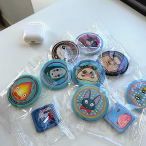 May include: A selection of cartoon character pin badges in clear packaging. The badges are round and square, with various designs and colours. An AirPod case is visible in the top left corner.