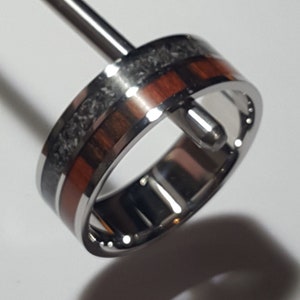 May include: A silver ring with a band of dark grey and red wood inlay. The ring is on a display stand.