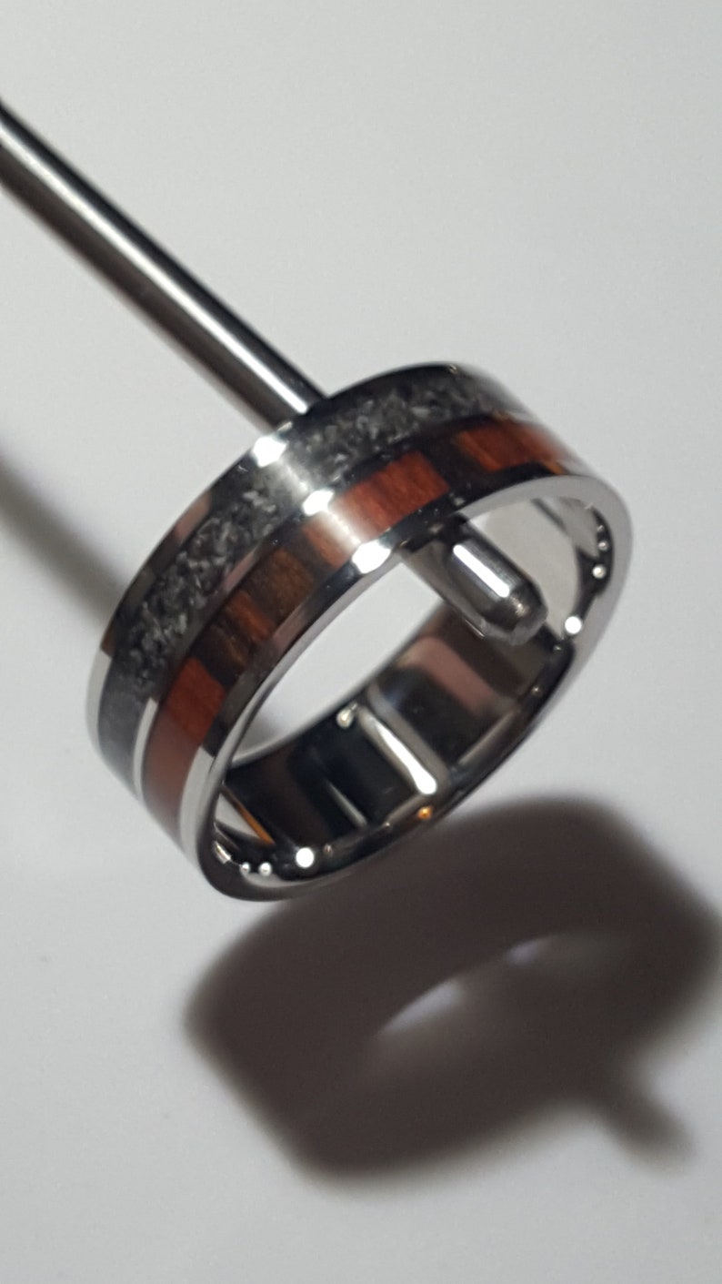 May include: A silver ring with a band of dark grey and red wood inlay. The ring is on a display stand.