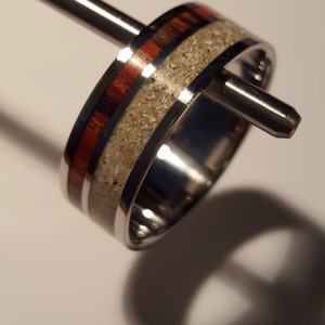 May include: A silver ring with a band of wood and white ash embedded in the metal. The ring is 8mm wide and is shown on a ring sizer.