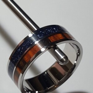 May include: A stainless steel ring with a blue and brown inlay. The ring is on a display stand.