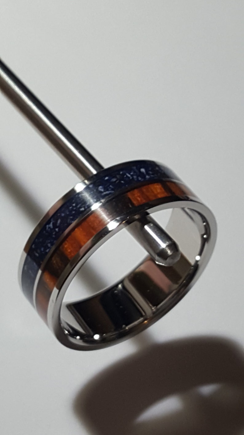 May include: A stainless steel ring with a blue and brown inlay. The ring is on a display stand.