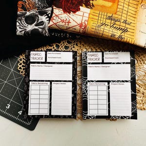May include: Two black and white fabric tracker notepads with spiderweb designs. The notepads have sections for fabric details and project notes. A black cutting mat and fabric with a skull print are in the background.