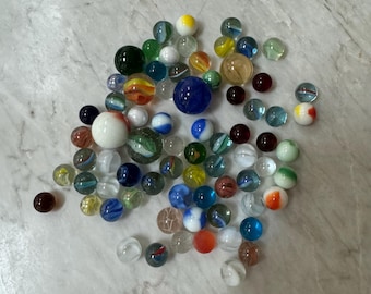 May include: A collection of colorful glass marbles in various shades of blue, green, red, yellow, and white. The marbles are arranged in a random pattern on a white marble surface.