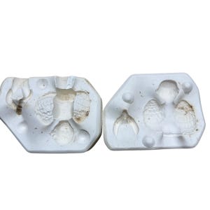 Puede incluir: Dos moldes de cerámica blanca para hacer una pequeña figura de ángel. Los moldes están abiertos y muestran los detalles de las alas, el cuerpo y la cara del ángel.
