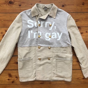 May include: A beige and gray jacket with a double breasted closure and gold buttons. The jacket has a gray panel on the front with the text "Sorry, I'm gay" printed in white.
