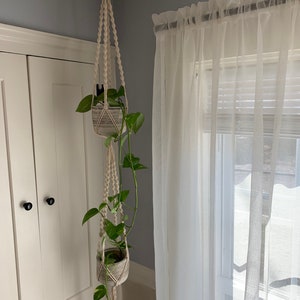 May include: A macrame plant hanger with two hanging pots, each containing a potted green plant. The hanger is made of natural cotton rope and is hanging from a ceiling hook. The pots are white and have a textured finish. The plants are trailing down the sides of the pots.