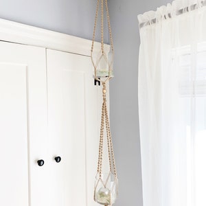 Puede incluir: Un colgador de plantas de macramé de dos niveles hecho de cuerda de color natural, suspendido del techo. Cada nivel contiene un jarrón de vidrio transparente, adecuado para plantas pequeñas o artículos decorativos. El fondo incluye una puerta blanca y una cortina transparente.