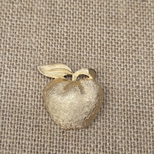 Puede incluir: Broche dorado en forma de manzana con superficie texturizada y un pequeño detalle de hoja. El broche se encuentra sobre un fondo neutro y texturizado, resaltando su acabado metálico y diseño clásico.