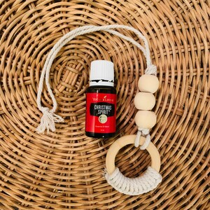Könnte beinhalten: Ein weißer Auto-Lufterfrischer aus Schnur mit Holzperlen und einem Ring. Neben dem Lufterfrischer befindet sich eine kleine Flasche Young Living Christmas Spirit ätherisches Öl.