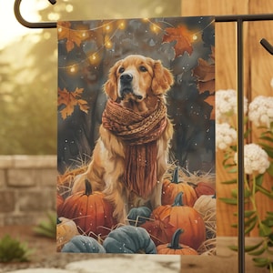 May include: A garden flag featuring a Golden Retriever wearing a scarf, surrounded by pumpkins and autumn leaves. The dog is the focal point, with a warm color palette of orange, brown, and green. Decorative lights are strung in the background.