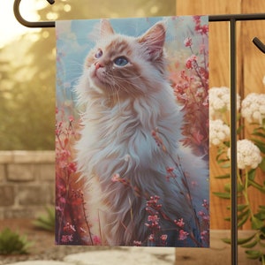 May include: A garden flag featuring a white and light brown cat with blue eyes, looking up, surrounded by pink flowers. The flag is hanging from a black metal pole.