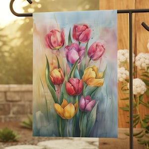 May include: A garden flag featuring a watercolor painting of tulips in shades of red, pink, and yellow. The flag is displayed on a black metal stand in an outdoor setting, with a blurred background of greenery and a stone pathway.