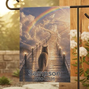 May include: A garden flag featuring a cat walking on a wooden bridge towards a rainbow. The flag has the name "Sampson" and the years "2010-2026" printed on it. The flag is displayed on a black metal stand in a garden setting.