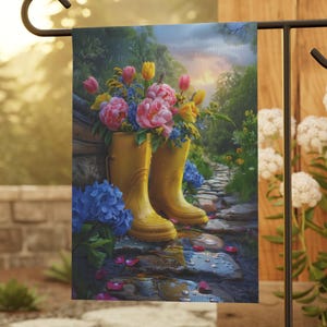 May include: A garden flag features a painting of yellow rain boots filled with pink and yellow flowers. The boots sit on a stone path with blue hydrangeas and a lush green background. The flag is displayed on a black metal stand.