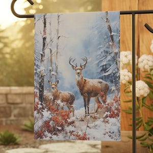 May include: A rectangular garden flag featuring a winter forest scene with two deer standing in snow among bare trees and red foliage. The flag is suspended from a black metal stand, displaying a detailed illustration of the animals in a cold landscape.