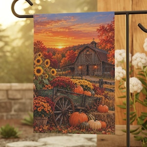 May include: A rectangular garden flag featuring a printed autumn scene of a rustic wooden barn at sunset, a weathered wagon filled with orange pumpkins and colorful fall flowers, and tall sunflowers standing in the foreground.