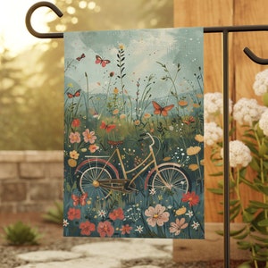 May include: A garden flag with a vintage bicycle illustration surrounded by colorful flowers and butterflies. The flag is attached to a black metal garden flag stand.
