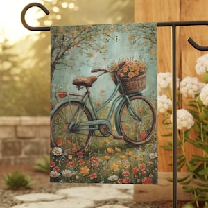 May include: A garden flag featuring a vintage bicycle with a basket full of flowers. The bicycle is painted a light blue color and has a brown wicker basket. The background is a soft blue with a floral pattern.