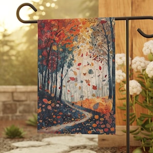 May include: A garden flag featuring a watercolor painting of an autumn forest scene. The artwork depicts a path winding through trees with colorful foliage in shades of red, orange, and yellow. The flag is displayed on a black metal stand.