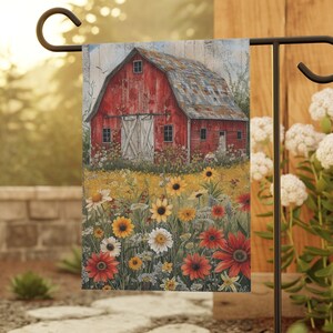 May include: A garden flag featuring a vibrant painting of a red barn with white doors, set amidst a field of colorful sunflowers and wildflowers. The flag is displayed on a black metal stand, perfect for outdoor decor.