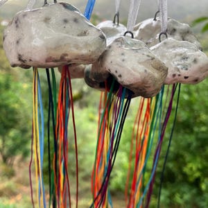 Può includere: Una collezione di ornamenti a forma di medusa in ceramica fatti a mano. Ogni medusa ha un corpo bianco sporco maculato con fili colorati pendenti. Gli ornamenti sono sospesi da ganci e nastri in metallo argentato, su uno sfondo verde sfocato.