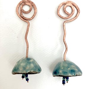 Può includere: Un paio di orecchini fatti a mano con un design in filo di rame. Ogni orecchino ha una parte superiore a spirale, un gambo sottile e un fondo a forma di campana in ceramica blu-verde con perline blu pendenti.