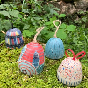 Puede incluir: Cuatro adornos de campana de cerámica pintados a mano con diseños únicos. Uno es azul con rayas verticales, otro rojo con detalles azules, un tercero azul con patrones texturizados y el último blanco con círculos rojos y azules.