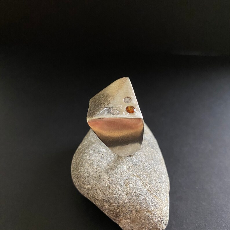 May include: A silver ring with a triangular face set with three gemstones. Two of the gemstones are white and one is orange.