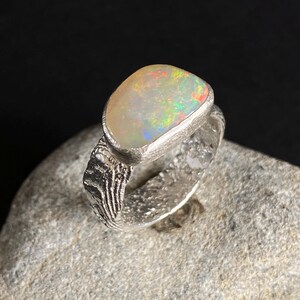 Könnte beinhalten: Ein Silberring mit einem großen, ovalen Opal-Edelstein. Der Opal zeigt ein lebendiges Farbspiel mit grünen, roten und gelben Blitzen. Der Ring hat ein strukturiertes Band und eine Lünettenfassung.
