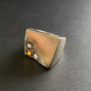 May include: A silver ring with a geometric design and three small gemstones: one orange, one white, and one clear.