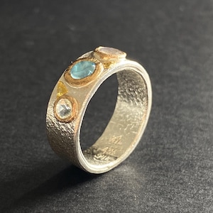 Könnte beinhalten: Ein silberner Ring mit strukturiertem Band, mit einem ovalen hellblauen Edelstein, einem runden, klaren Edelstein, einem kleinen gelben Edelstein und einem kleinen hellvioletten Edelstein. Die Edelsteine sind in Goldfassungen gefasst.