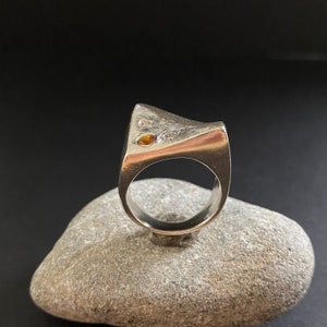 May include: A silver ring with a unique, angular design. The ring features two small, round gemstones, one orange and one clear.