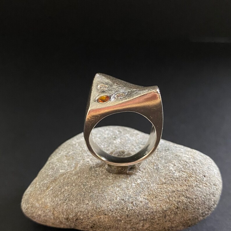 May include: A silver ring with a unique, angular design. The ring features two small, round gemstones, one orange and one clear.