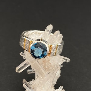 Könnte beinhalten: Ein silberner Ring mit einem blauen Edelstein und goldenen Akzenten. Der Ring liegt auf einem Bett aus weißen Kristallen.