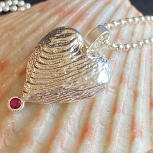 Può includere: Un ciondolo a forma di cuore in argento con una superficie testurizzata e striata che ricorda una conchiglia, sospeso a una catenina a palline in argento. Una piccola pietra preziosa rotonda rosso scuro è incastonata sulla punta del cuore.