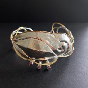 May include: Silver cuff bracelet with a hammered leaf design. The bracelet has a gold-toned wire frame and three small, round, red gemstones. The leaf has a textured surface and a swirling detail.