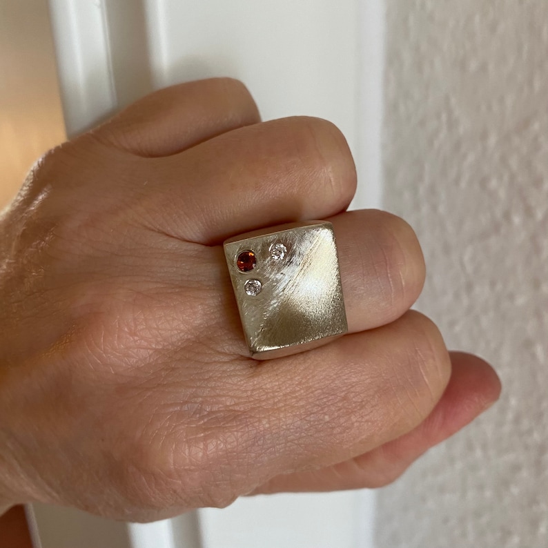 May include: A silver square ring with a brushed finish and three gemstones. Two of the gemstones are small and clear, and one is larger and red.
