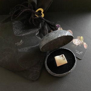 May include: A silver ring with a square face and three small red gemstones set in the center. The ring is displayed in a black velvet box with a gray stone exterior. The box is open and the ring is visible inside. The box is sitting on a black surface.