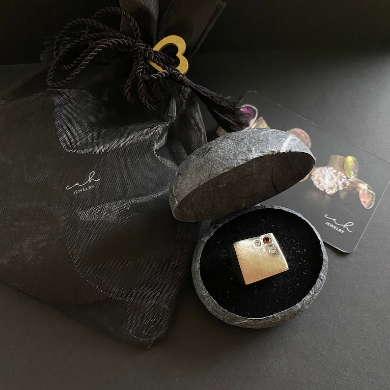 May include: A silver ring with a square face and three small red gemstones set in the center. The ring is displayed in a black velvet box with a gray stone exterior. The box is open and the ring is visible inside. The box is sitting on a black surface.