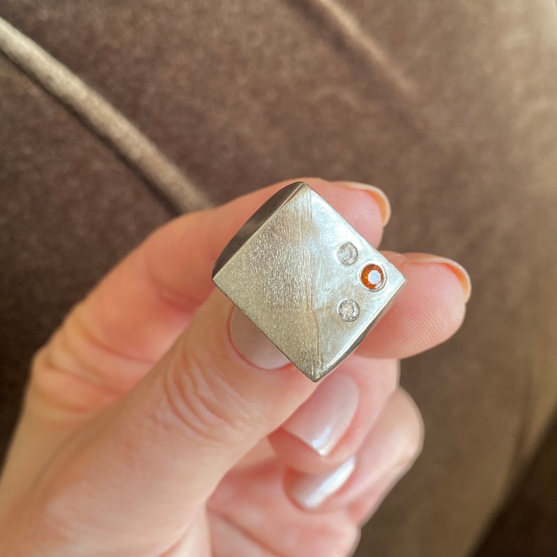 May include: A silver square ring with three small gemstones. Two of the gemstones are clear and one is orange.