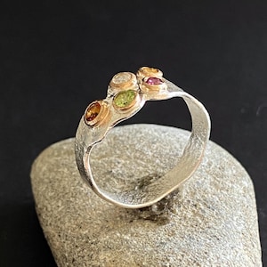 Könnte beinhalten: Ein Silberring mit fünf farbigen Edelsteinen. Die Edelsteine sind in Goldfassungen gefasst und umfassen orangefarbene, grüne, klare und rosa Steine. Der Ring wird auf einem grauen Stein vor schwarzem Hintergrund präsentiert.