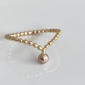 Op de afbeelding: Een goudkleurige ring met een kralenband en een hangende, ronde, roségoudkleurige bedel. De ring heeft een V-vormig ontwerp met de bedel die aan de punt hangt. De ring ligt op een wit oppervlak.